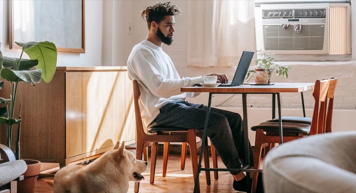 A man working at a table with a laptop and papers while his dog lies on the wooden floor beside him